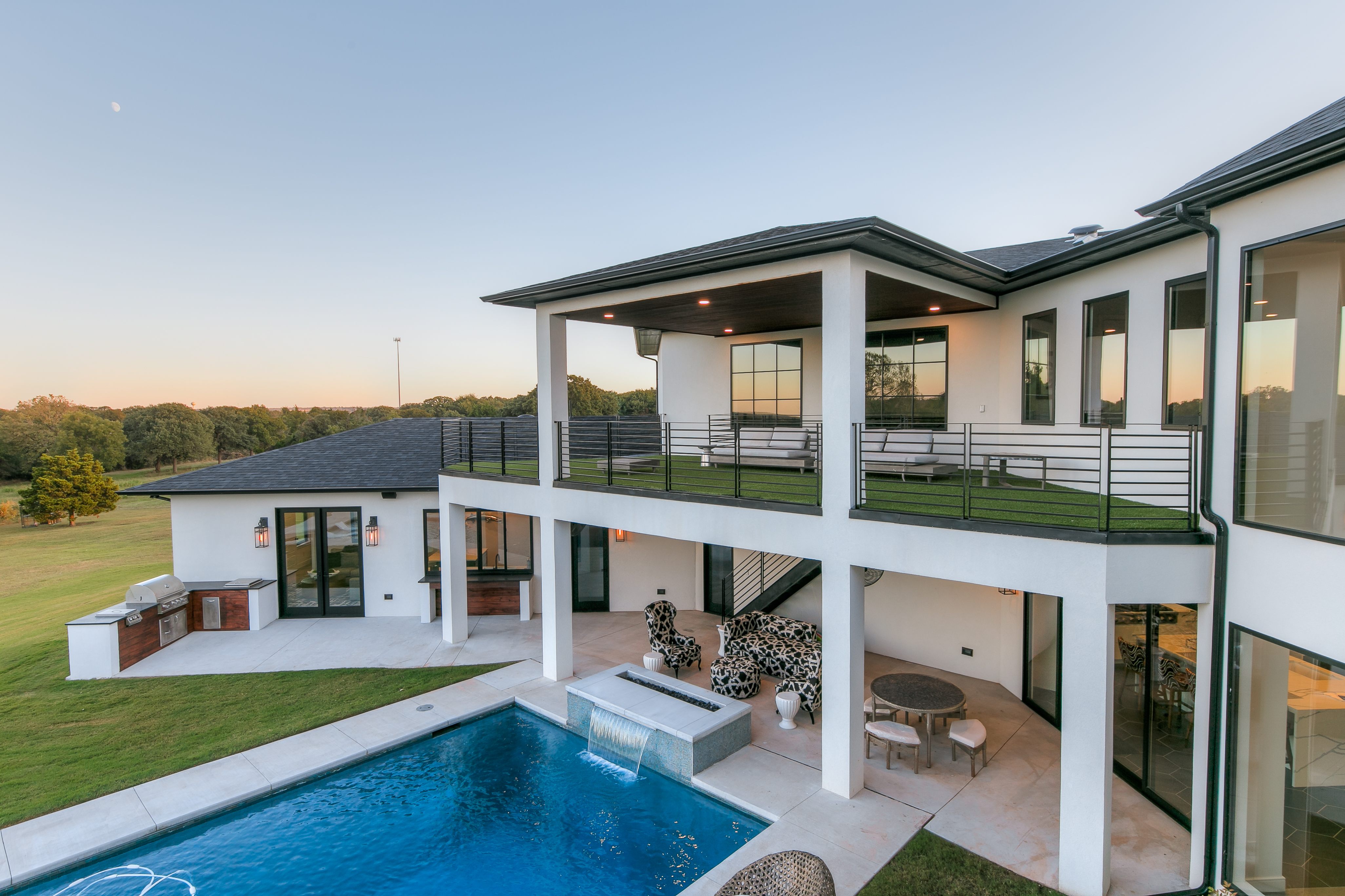Modern two-story custom home at dusk with covered balcony, outdoor living, and pool with water feature